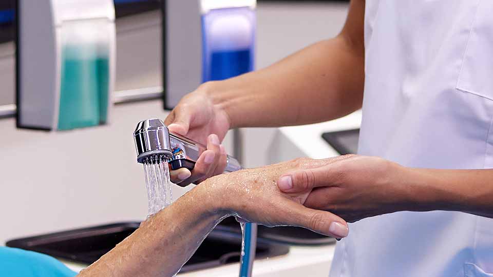 Mobile Bettdusche MoDu im Einsatz. Duschkopf sprüht Wasser auf Unterarm von Patientin.